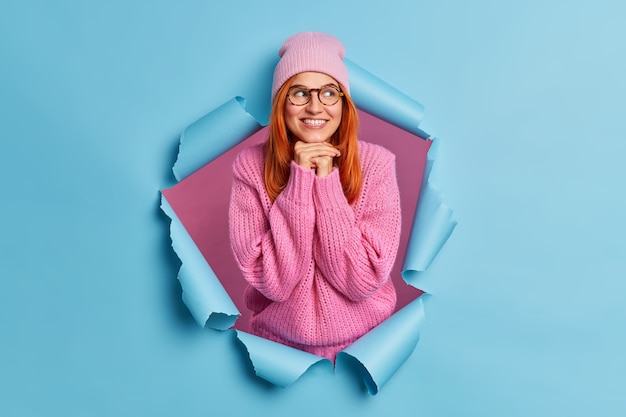 Heureux Jeune Femme Rousse Garde Les Mains Sous Le Menton Sourit Agréablement Et Regarde Ailleurs Pense à Quelque Chose De Très Bon Ou D'agréable Porte Des Lunettes De Chapeau De Chandail D'hiver.