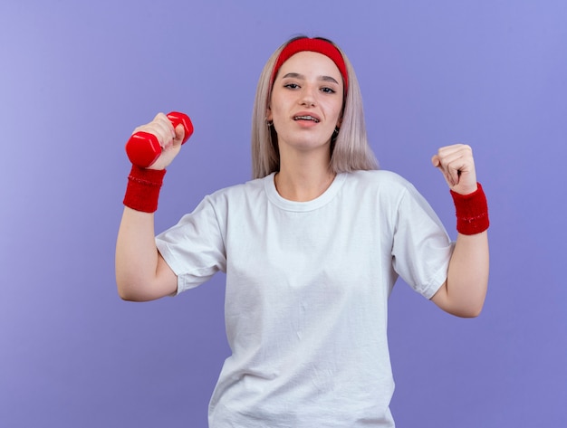 Heureux jeune femme sportive avec des accolades portant un bandeau et des bracelets garde le poing et détient un haltère isolé sur un mur violet