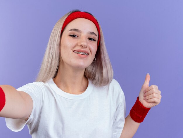 Heureux jeune femme sportive avec des accolades portant bandeau et bracelets Thumbs up et regarde à l'avant isolé sur mur violet