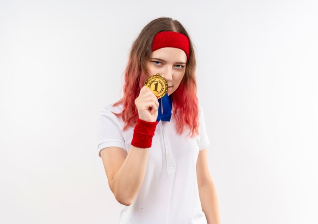 Heureux jeune femme sportive en bandeau montrant sa médaille d'or à la confiance debout sur un mur blanc