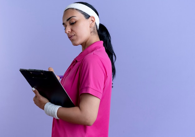 Heureux jeune femme sportive caucasienne portant bandeau et bracelets se tient sur le côté et écrit avec un stylo dans le presse-papiers sur violet avec copie espace