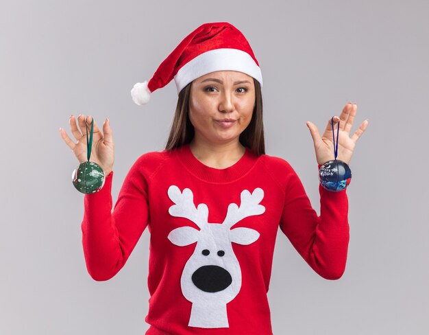 Heureux jeune fille asiatique portant chapeau de Noël avec chandail tenant des boules de sapin de Noël isolé sur fond blanc