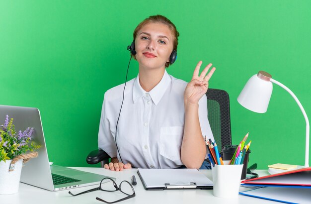Heureux jeune fille blonde du centre d'appels portant un casque assis au bureau avec des outils de travail montrant trois avec la main