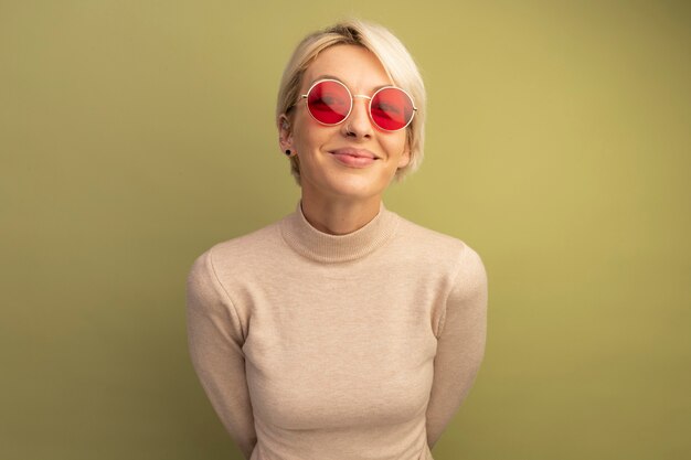 Heureux jeune fille blonde portant des lunettes de soleil en gardant les mains derrière le dos