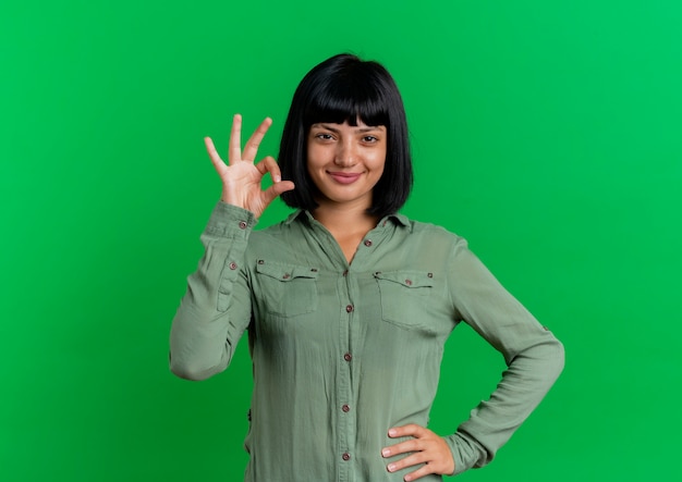 Heureux jeune fille brune caucasienne met la main sur la taille gesticulant signe de la main ok isolé sur fond vert avec espace copie