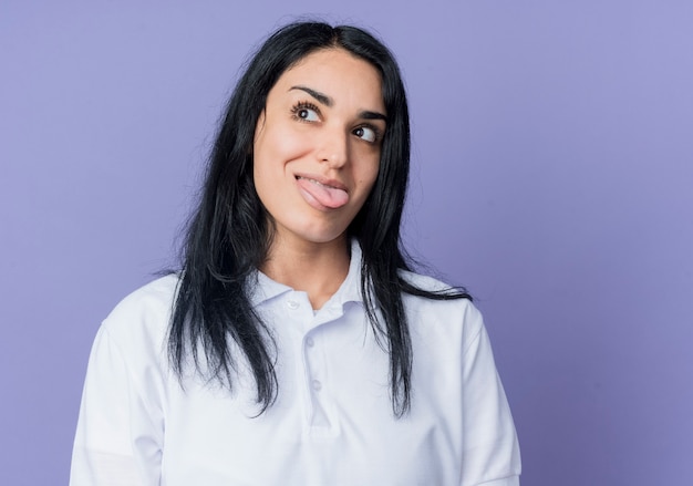 Heureux jeune fille caucasienne brune sort la langue à côté isolé sur mur violet