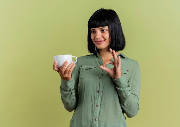 Photo gratuite heureux jeune fille caucasienne brune tient la tasse et les gestes ok signe de la main regardant sur le côté