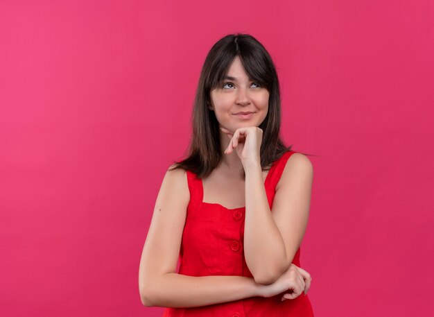 Heureux jeune fille caucasienne mettant la main sur le menton et levant les yeux sur fond rose isolé avec copie espace