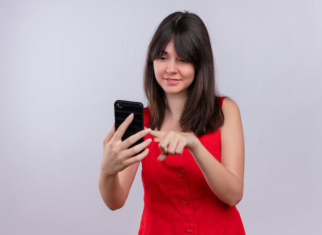 Heureux jeune fille caucasienne tenant le téléphone et mettre le doigt sur l'écran en regardant le téléphone sur fond blanc isolé avec copie espace