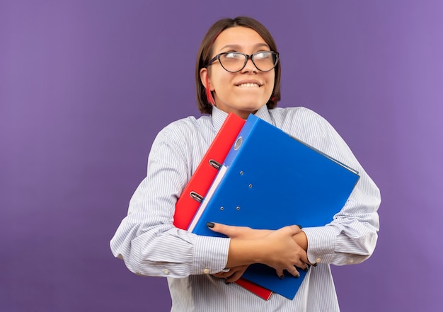 Heureux jeune fille de centre d'appels portant des lunettes tenant des dossiers à côté isolé sur fond violet avec copie espace