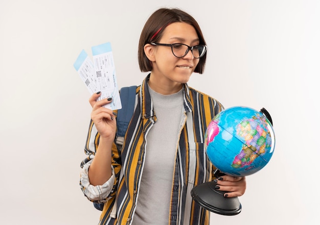 Heureux jeune fille étudiante portant des lunettes et sac à dos tenant des billets d'avion et globe regardant globe isolé sur mur blanc