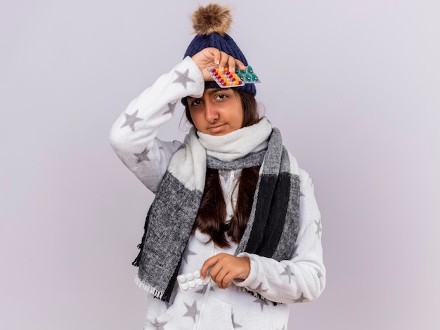 Heureux jeune fille malade portant chapeau d'hiver avec foulard tenant des pilules mettant la main sur le front isolé sur fond blanc