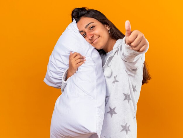 Heureux jeune fille malade regardant la caméra hugging oreiller montrant le pouce vers le haut isolé sur jaune