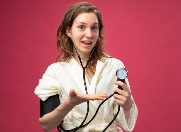 Heureux jeune fille malade regardant droit devant portant une robe blanche mesurant sa propre pression avec sphygmomanomètre isolé sur rose