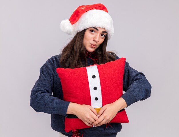 Heureux jeune fille de race blanche avec Bonnet de Noel et guirlande autour du cou tient un oreiller décoré à la recherche