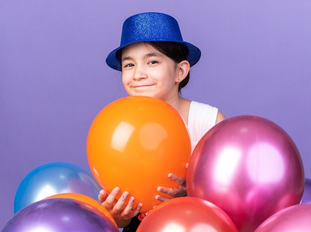 Heureux jeune fille de race blanche avec chapeau de fête bleu debout avec des ballons d'hélium isolés sur un mur violet avec espace de copie