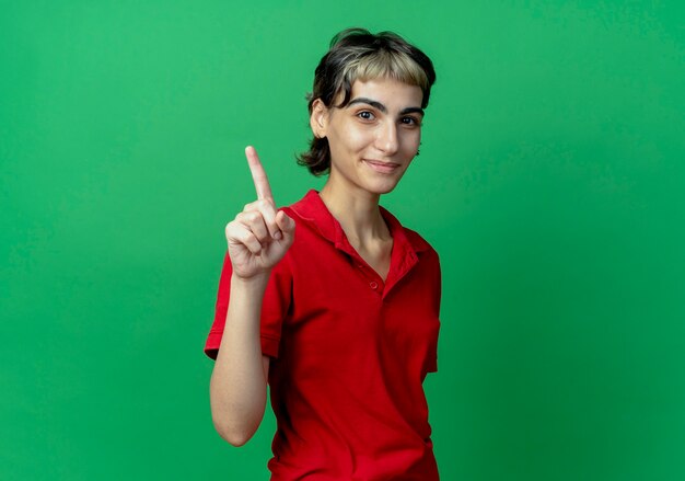 Heureux jeune fille de race blanche avec coupe de cheveux de lutin levant le doigt isolé sur fond vert avec espace de copie