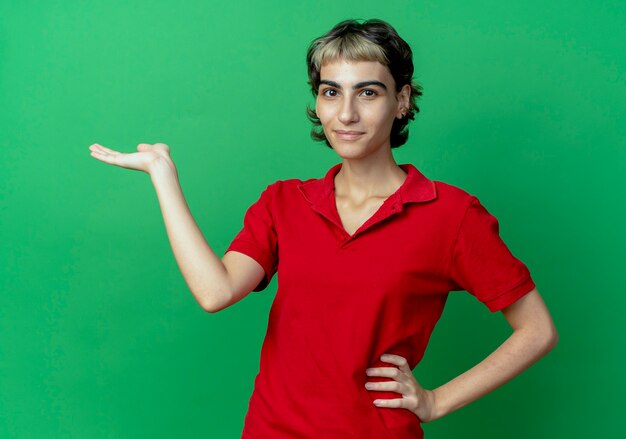 Heureux jeune fille de race blanche avec coupe de cheveux de lutin mettant la main sur la taille et montrant la main vide isolée sur fond vert