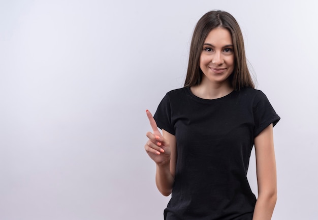 Heureux jeune fille de race blanche portant un t-shirt noir pointe le doigt vers le haut sur un mur blanc isolé