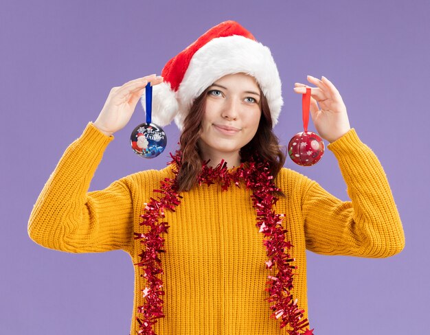 Heureux jeune fille slave avec Bonnet de Noel et avec guirlande autour du cou tenant des ornements de boule de verre isolés sur fond violet avec espace copie