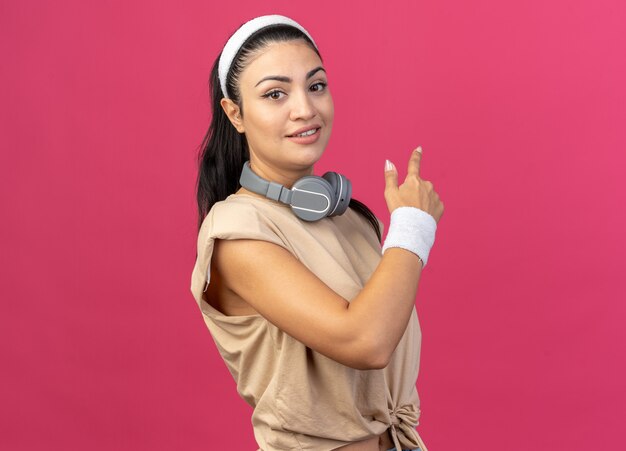 Heureux jeune fille sportive caucasienne portant un bandeau et des bracelets avec des écouteurs autour du cou, debout dans la vue de profil regardant l'avant pointant derrière isolé sur un mur rose