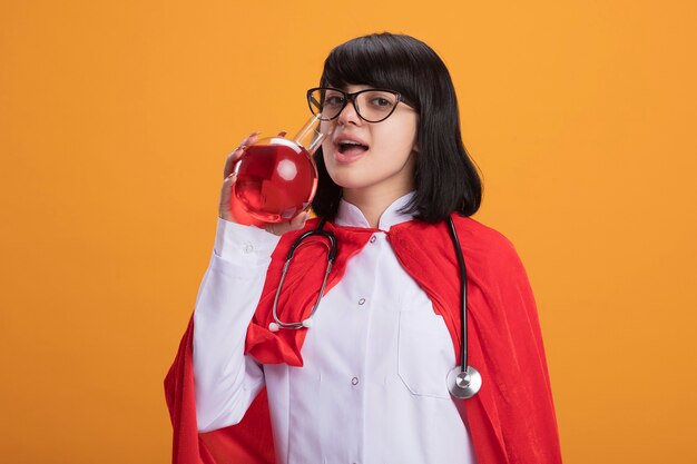 Heureux jeune fille de super-héros portant un stéthoscope avec une robe médicale et une cape avec des verres boit un liquide rouge à partir de verre de chimie