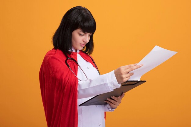 Heureux jeune fille de super-héros portant un stéthoscope avec robe médicale et manteau feuilletant le presse-papiers isolé sur mur orange