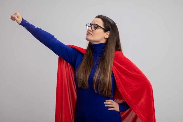 Heureux jeune fille de super-héros regardant côté portant des lunettes levant la main isolé sur fond blanc