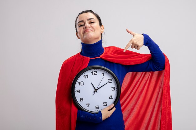 Heureux jeune fille de super-héros tenant et points à l'horloge murale isolé sur fond blanc