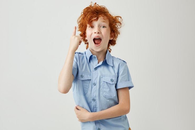Photo gratuit heureux jeune garçon aux cheveux roux bouclés et taches de rousseur en chemise bleue pointant à l'envers avec une expression heureuse et excitée