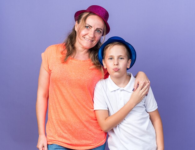 Heureux jeune garçon slave avec chapeau de fête bleu debout avec sa mère portant chapeau de fête violet isolé sur mur violet avec espace copie