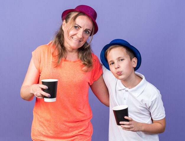 Heureux jeune garçon slave avec chapeau de fête bleu tenant une tasse de papier debout avec sa mère portant un chapeau de fête violet isolé sur mur violet avec espace copie