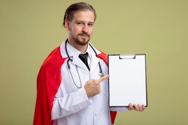 Heureux jeune gars de super-héros portant un stéthoscope avec une robe médicale tenant et des points au presse-papiers isolé sur fond vert olive