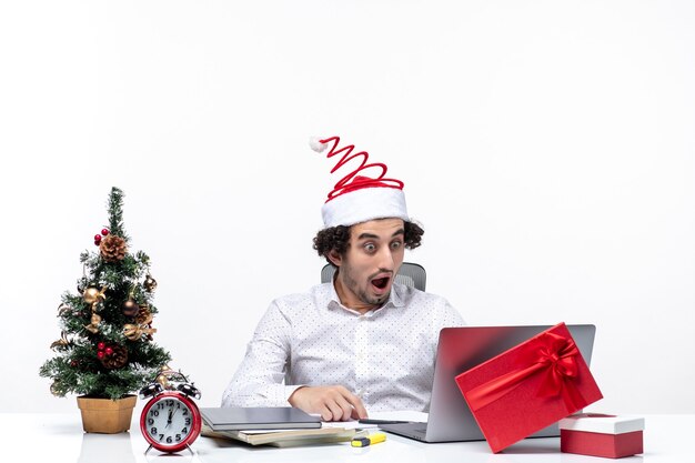 Heureux jeune homme d'affaires choqué positif avec drôle de chapeau de père Noël vérifiant soigneusement son au bureau sur fond blanc