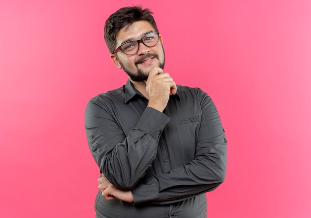 Heureux jeune homme d'affaires portant des lunettes mettant la main sous le menton isolé sur un mur rose