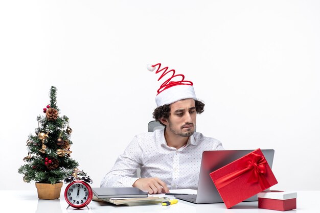 Heureux jeune homme d'affaires positif avec drôle de chapeau de père Noël vérifiant son soigneusement au bureau sur fond blanc