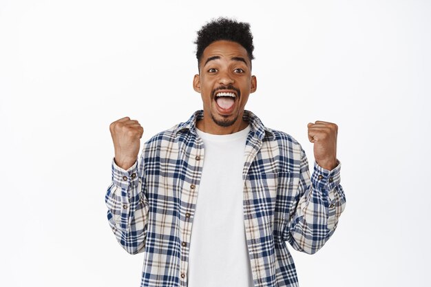 Heureux jeune homme afro-américain célébrant, gagnant, enracinant pour l'équipe, serrer et faire pomper le poing pour triompher gagner, atteindre l'objectif, regardant étonné la caméra, fond blanc.