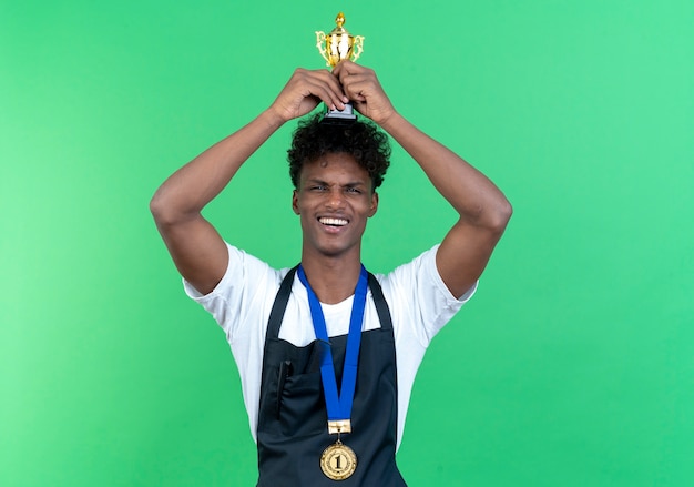 Photo gratuite heureux jeune homme afro-américain coiffeur portant l'uniforme et la médaille soulevant la coupe du vainqueur isolé sur fond vert