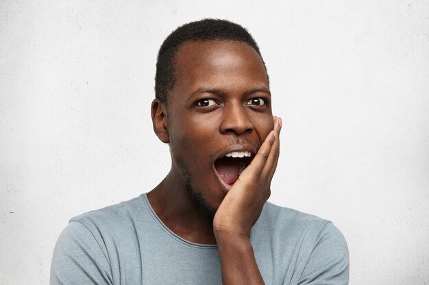 Heureux jeune homme afro-américain excité en t-shirt gris ouvrant la bouche avec étonnement, touchant son visage et regardant avec une expression fascinée, stupéfait par une excellente idée qui lui traversa l'esprit