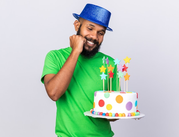 Heureux jeune homme afro-américain portant un chapeau de fête tenant un gâteau mettant la main sur la joue isolée sur un mur blanc