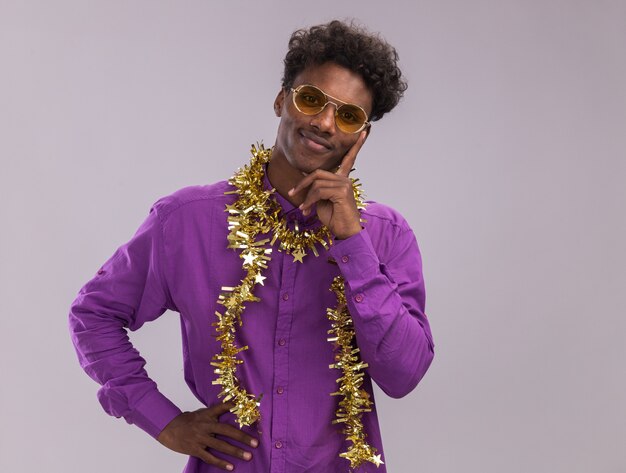 Heureux jeune homme afro-américain portant des lunettes avec une guirlande de guirlandes autour du cou regardant la caméra en gardant la main sur la taille et sur le menton isolé sur fond blanc avec espace de copie