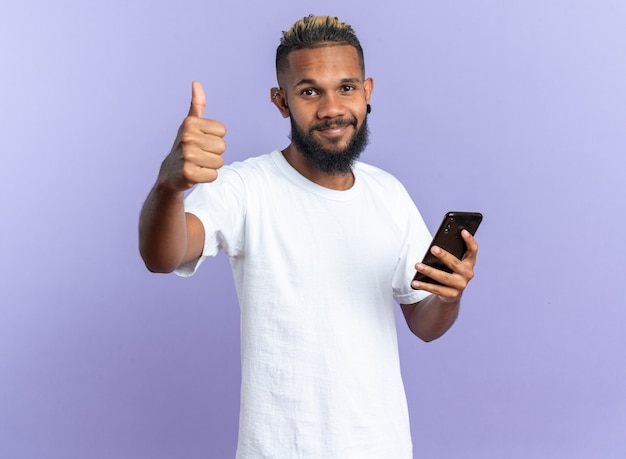 Heureux jeune homme afro-américain en t-shirt blanc tenant un smartphone regardant la caméra montrant les pouces vers le haut souriant joyeusement