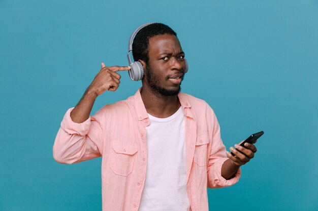 Heureux jeune homme afro-américain tenant un téléphone portant des écouteurs isolés sur fond bleu