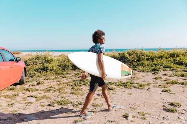 Heureux jeune homme afro-américain venant avec planche de surf le long de la côte