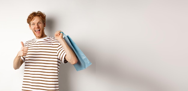 Photo gratuite heureux jeune homme aux cheveux rouges faisant du shopping dans les magasins montrant le pouce levé et tenant un sac en papier sur l'épaule