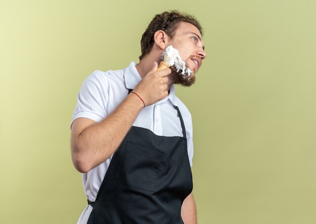 Heureux jeune homme barbier portant une crème à raser appliquée uniforme avec blaireau isolé sur mur vert olive