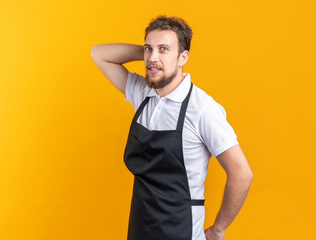 Heureux jeune homme barbier en uniforme mettant la main sur le cou isolé sur mur jaune