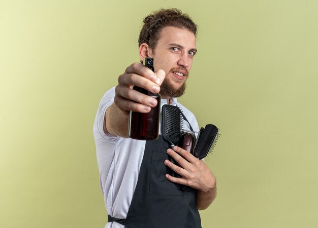 Heureux jeune homme barbier en uniforme tenant des outils de barbier et tenant un vaporisateur isolé sur un mur vert olive