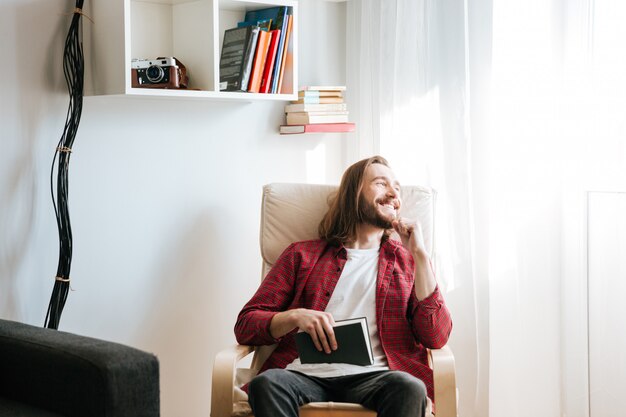 Heureux jeune homme barbu avec livre assis sur un fauteuil et regardant la fenêtre à la maison