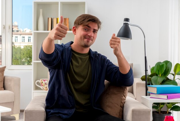 Heureux jeune homme beau blond est assis sur un fauteuil, les pouces vers le haut avec deux mains à l'intérieur du salon